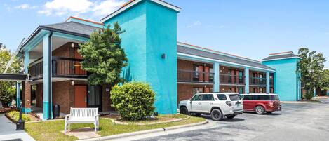 A hotel with a turquoise facade, a covered outdoor seating area, and a parking lot with two vehicles.