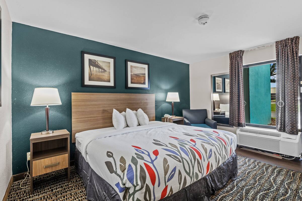 A hotel room with a bed, two bedside lamps, a nightstand, a chair, a window with curtains, and two framed pictures on the wall.