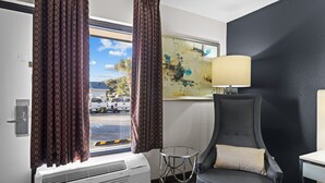 A hotel room with a large window, a chair, a table, and a wall painting. - Garnet Inn & Suites, Morehead City near Atlantic Beach (Morehead City)