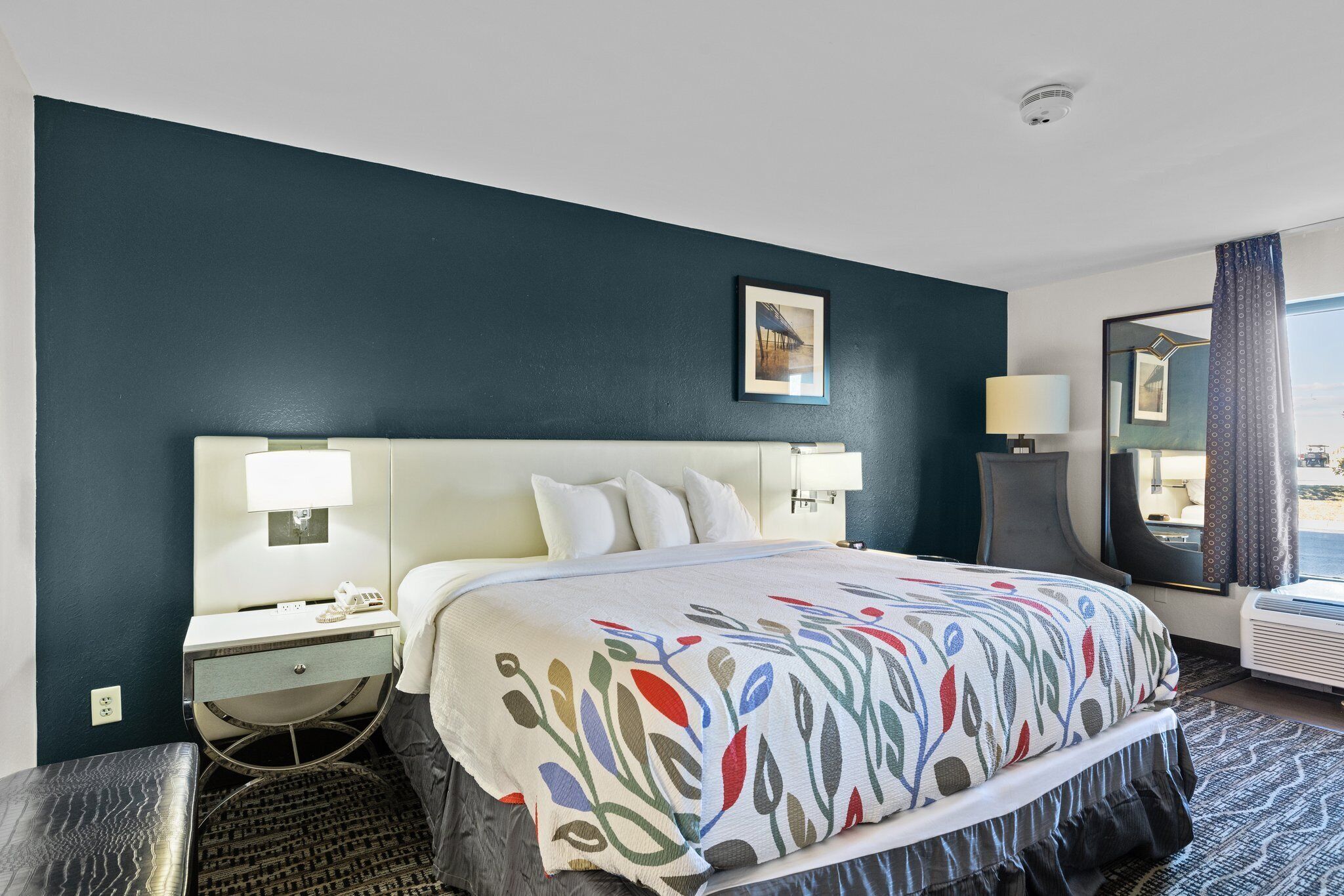 A hotel room with a large bed, two bedside tables with lamps, a small bench, a window with curtains, and a view of the outside.