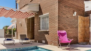 Piscina com degraus, uma cadeira de praia rosa e duas cadeiras de metal com almofadas.