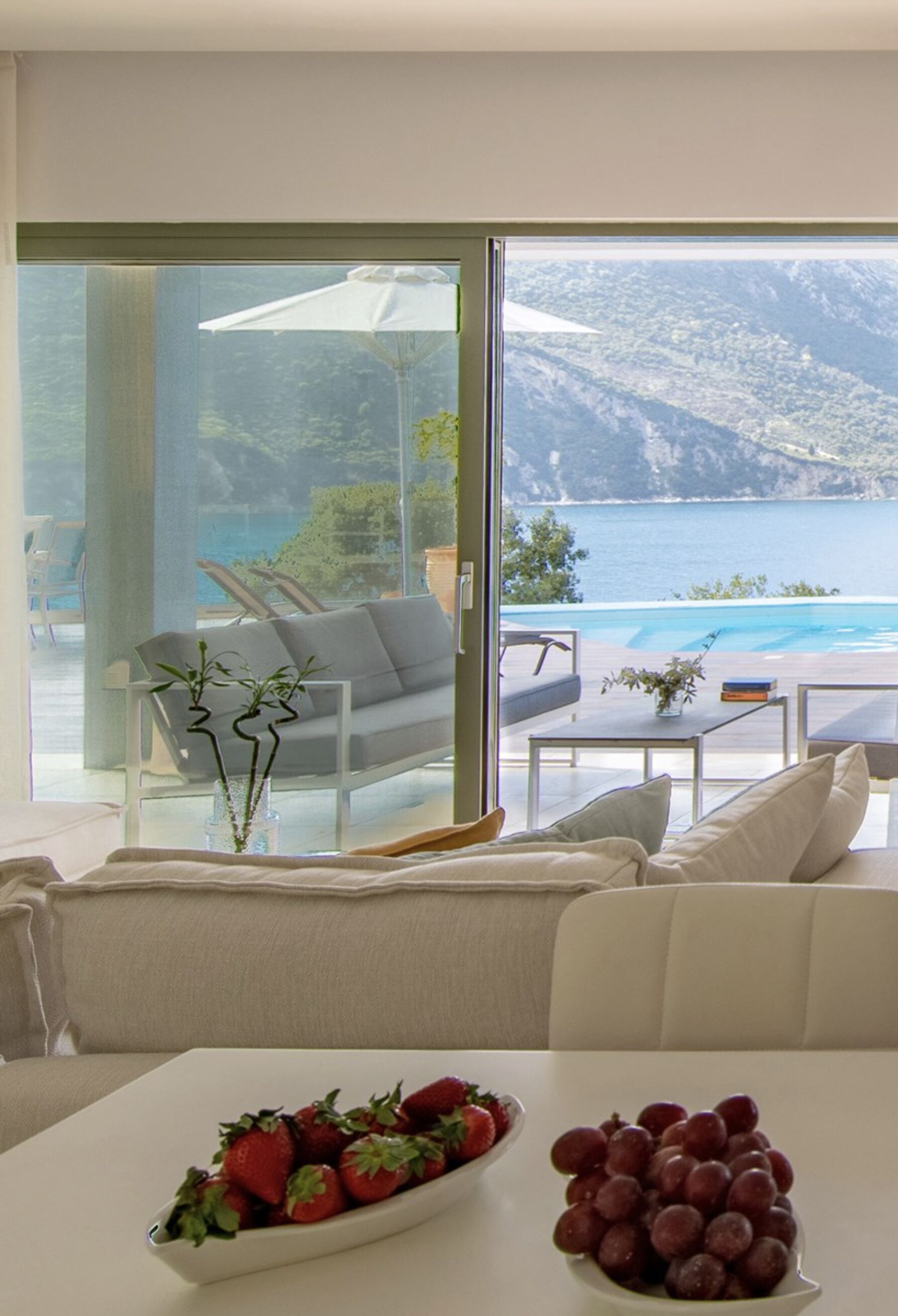 Una sala moderna con un gran ventanal que da a una piscina y al mar. Hay un sofá, una mesa de centro y un frutero en la mesa.