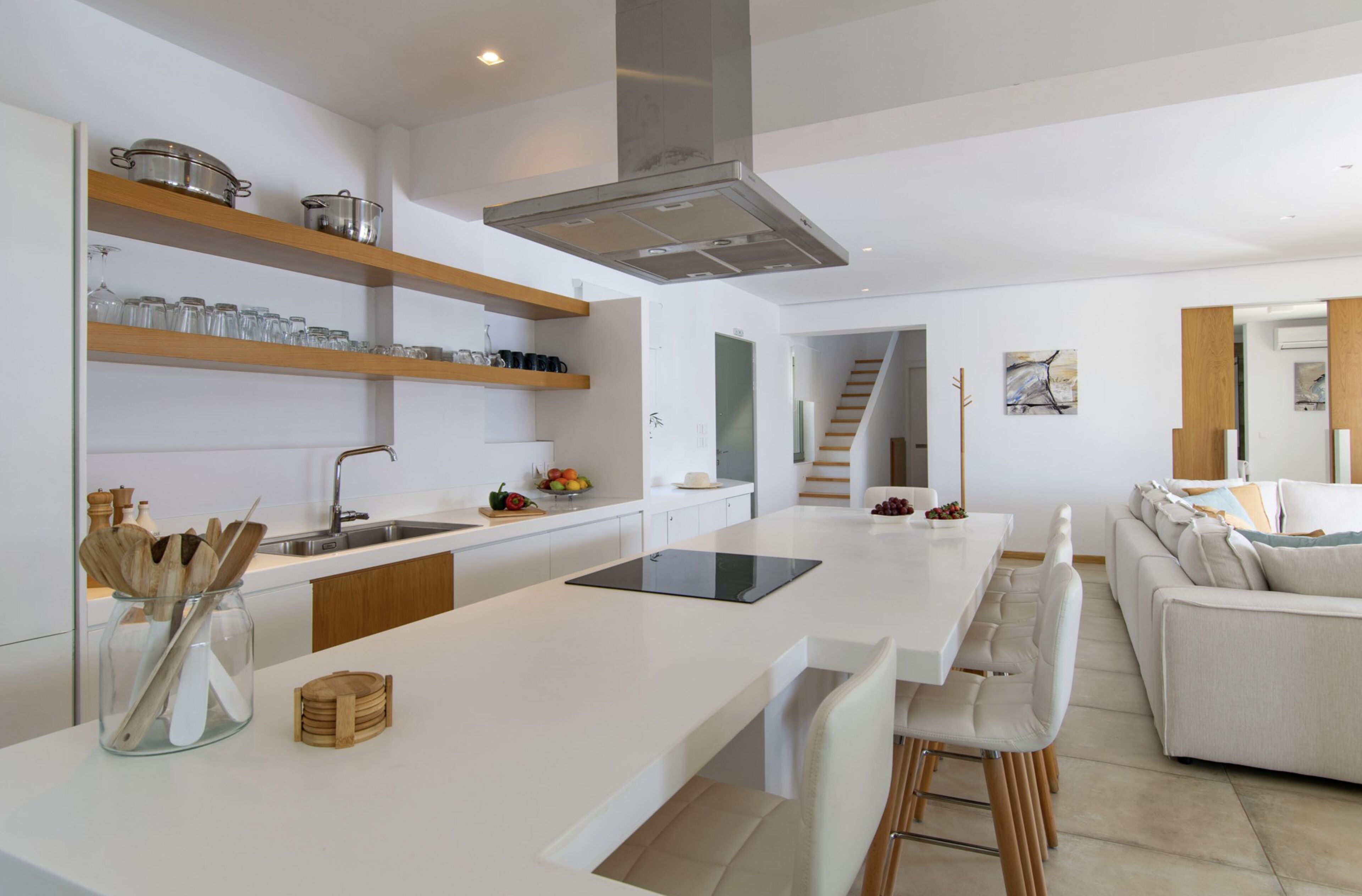 Une cuisine moderne avec des plans de travail blancs, des étagères en bois et un îlot central. Un escalier mène à un autre niveau.