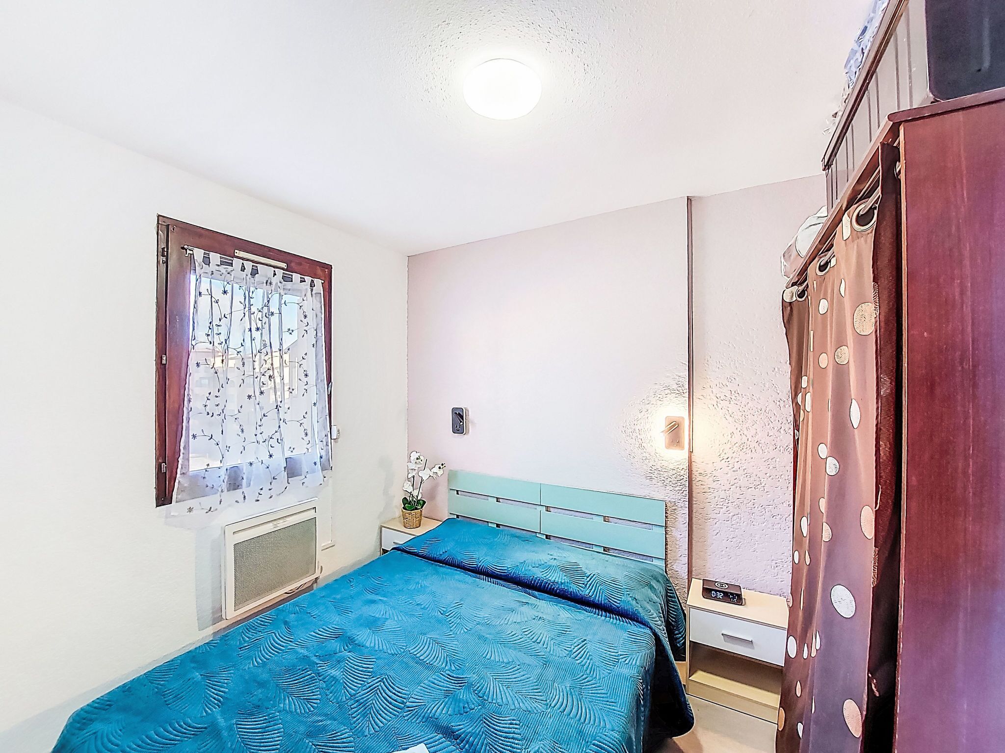 Une chambre à coucher comprenant un lit, une table de chevet, une horloge murale, une fenêtre avec un rideau et une armoire.