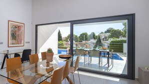 A modern dining area with a glass table and chairs, a cactus on the table, and a view of a swimming pool and greenery outside. - Bellveure by Interhome (Benissa)