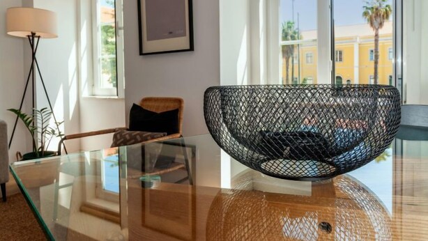 En moderne stue med sofabord i glas, en fletstol, en gulvlampe og et billede i ramme på væggen.