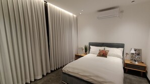 Un dormitorio moderno con una cama grande, cortinas blancas, un cabecero gris y una mesita de noche pequeña con una lámpara y un cartel.