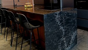 Una cocina moderna con una isla de mármol oscuro, taburetes negros, horno empotrado y refrigerador.