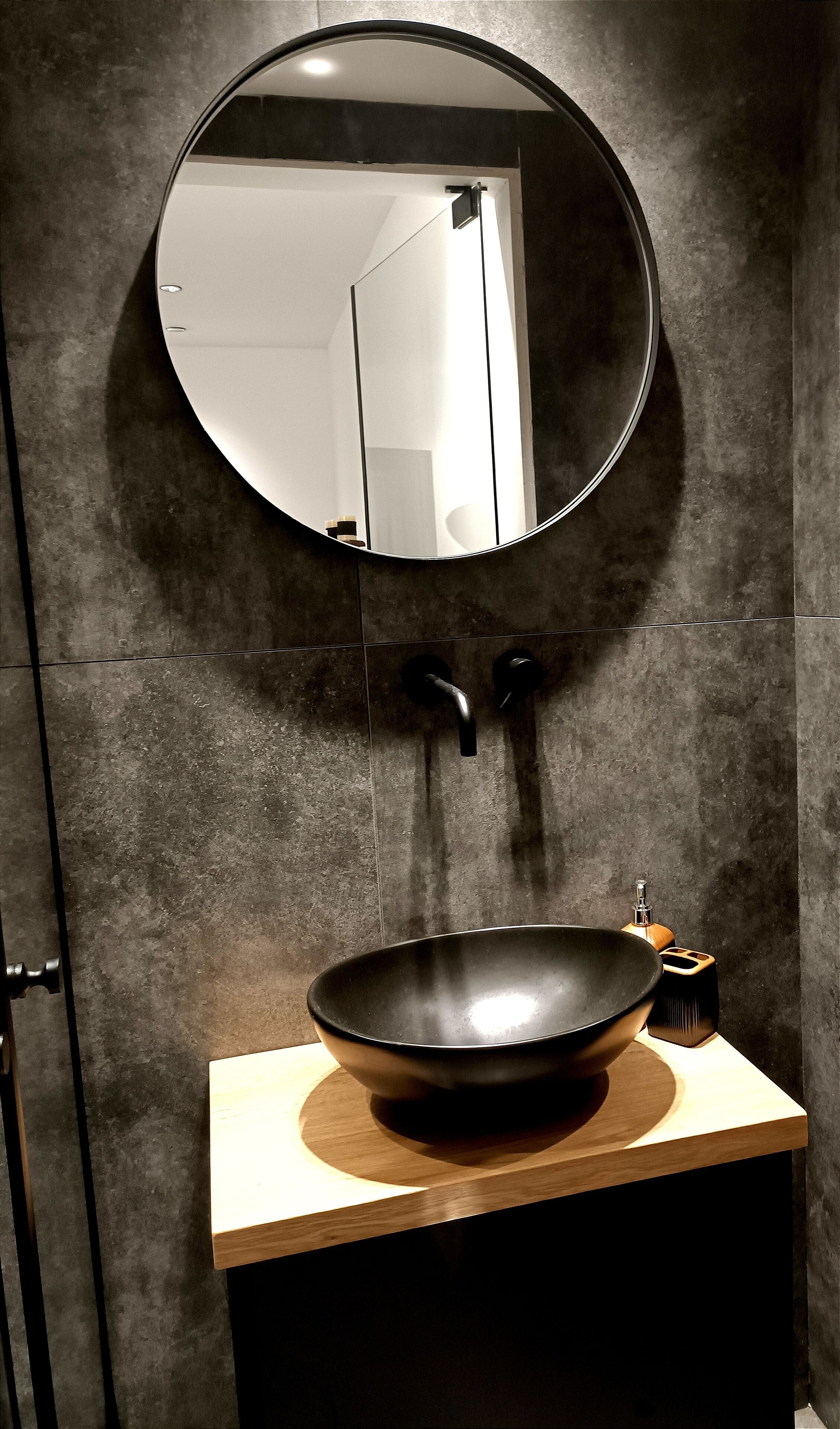 Un bany amb un mirall rodó, un lavabo negre i un armari de tocador de fusta.