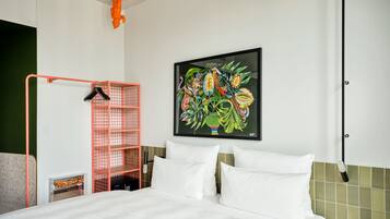 A hotel room with a bed, white pillows, a colorful artwork on the wall, a red storage unit, and a ceiling light fixture.