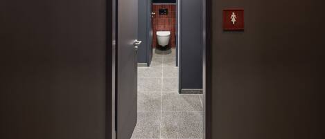 A hallway with a door leading to a bathroom. The bathroom has a toilet, a sink, and a shower area. There is a red emergency exit sign on the wall.
