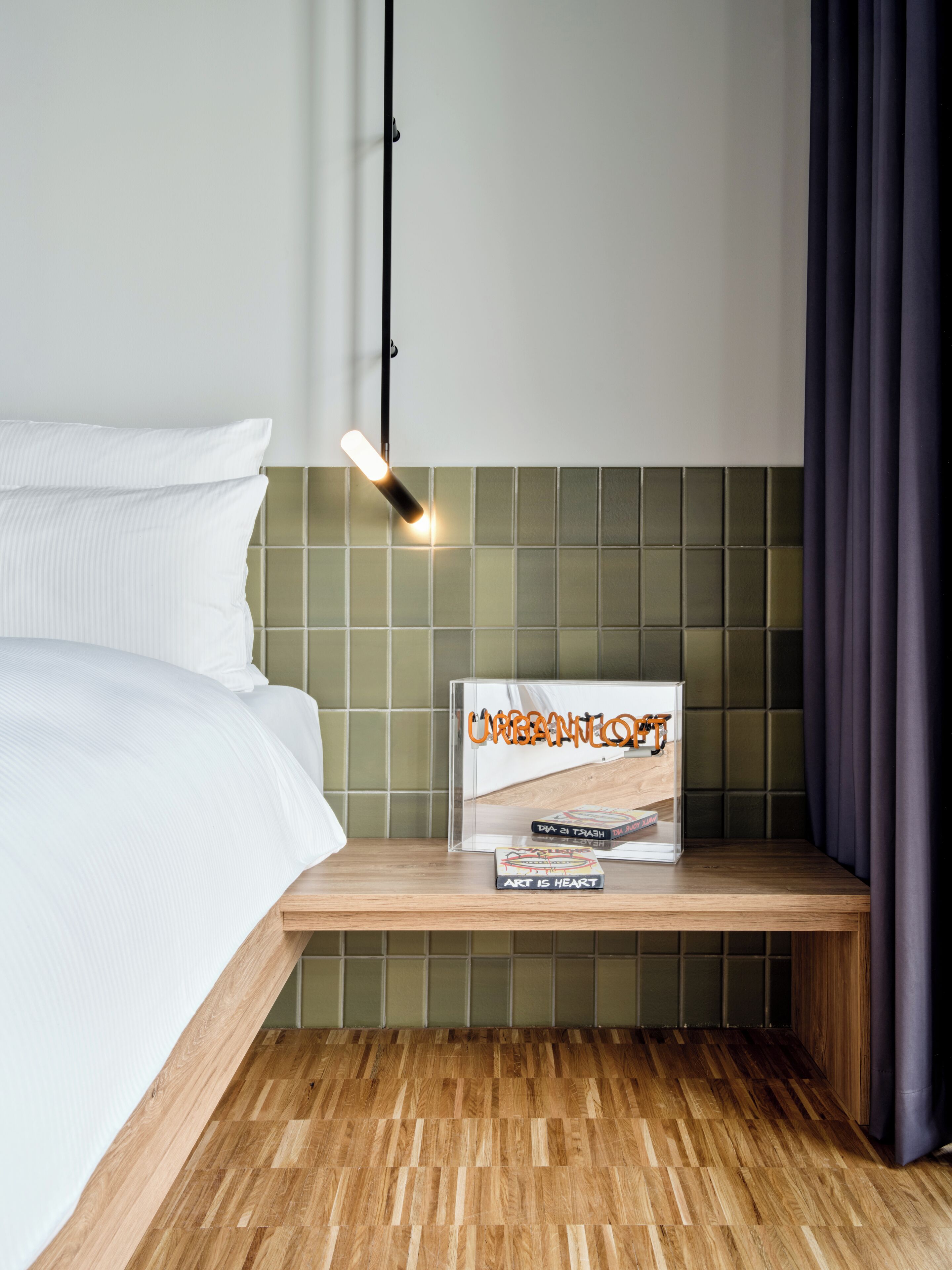 Una cama bien tendida con sábanas blancas, una mesita de noche con un libro titulado 'Urban Jungle' y una lámpara moderna en el techo.