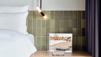 Una cama bien tendida con sábanas blancas, una mesita de noche con un libro titulado 'Urban Jungle' y una lámpara moderna en el techo.