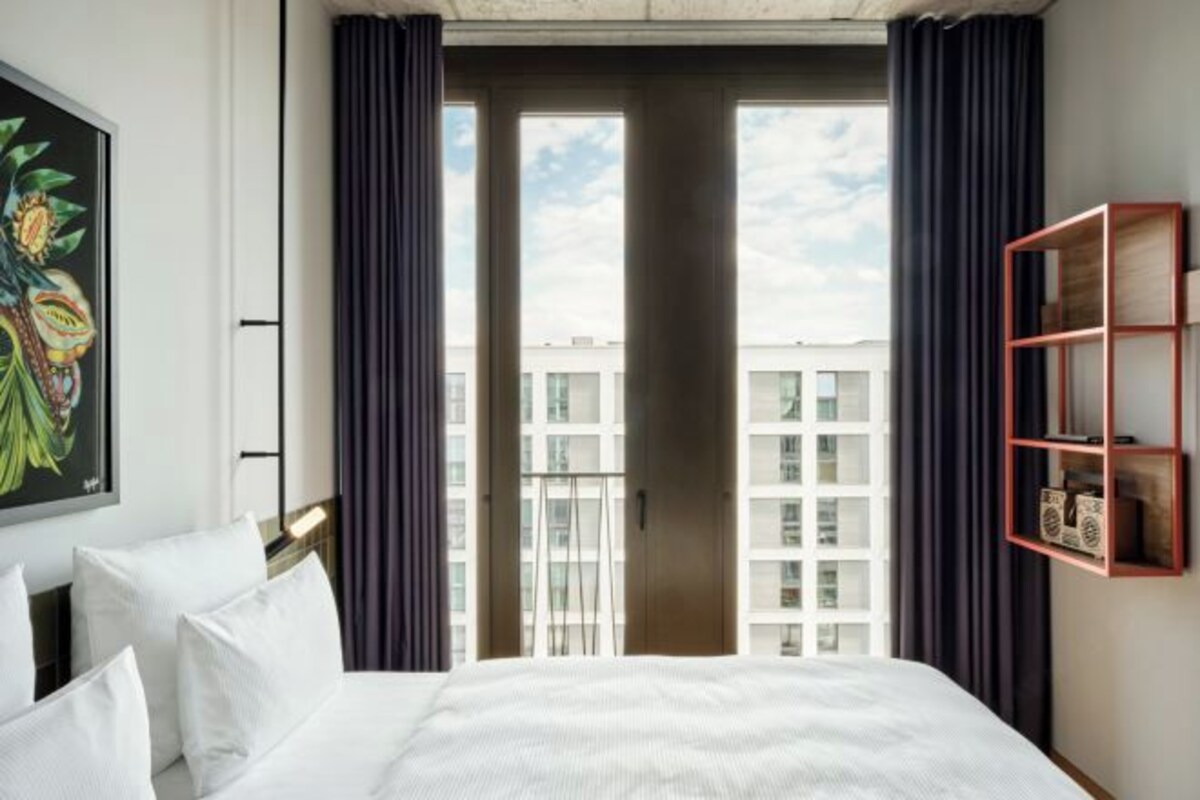 A hotel room with a large bed, a view of the city through a glass door, and a red-framed shelf with books.