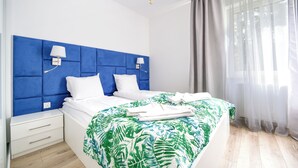 Une chambre à coucher moderne avec une tête de lit bleue, du linge de lit blanc et une couvre-lit ornée d’un motif de feuilles tropicales.
