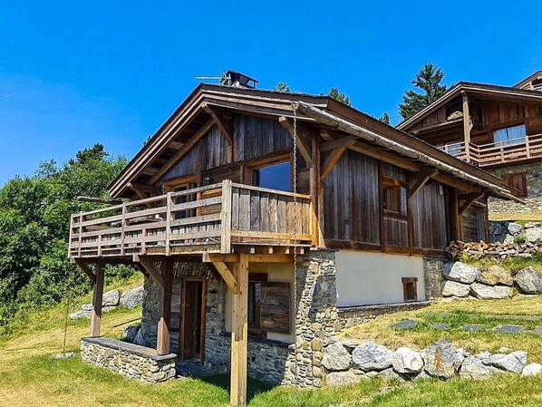 A wooden chalet with a balcony, stone foundation, and a stone wall.