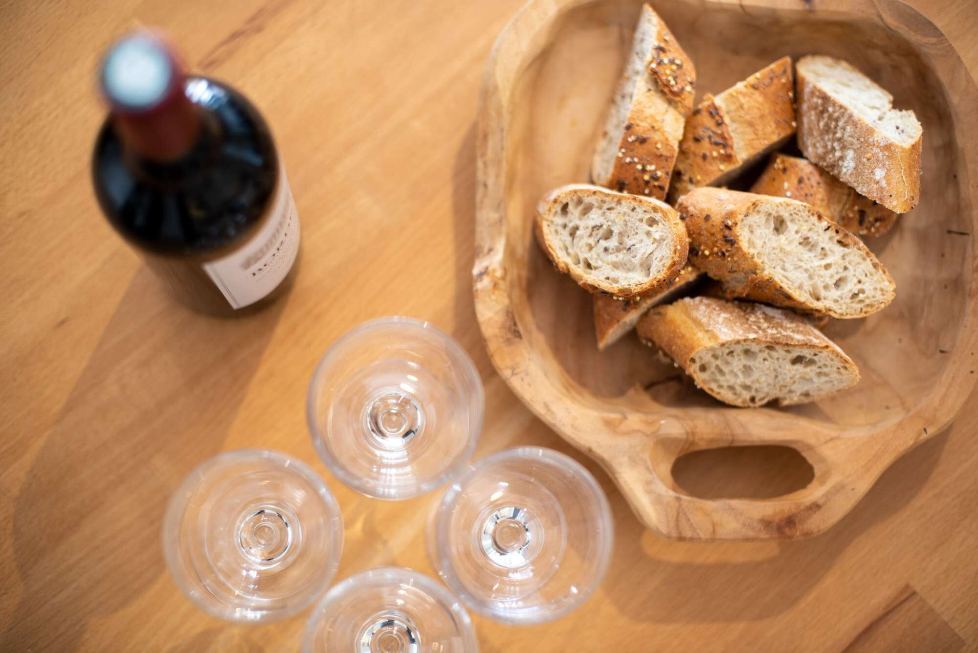 Una mesa de madera con un bol de pan en rodajas, una botella y cuatro copas de vino.