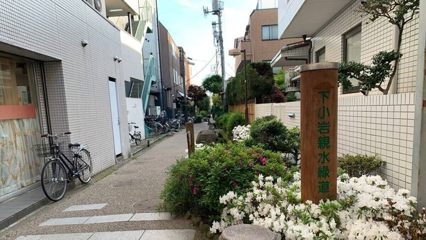 En smal byvej med række af cykler parkeret på venstre side, en stenpost med japanske tegn, og en lille have med hvide blomster.