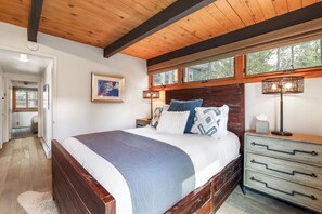 A bedroom with a large bed, wooden headboard, bedside tables, a nightstand, a lamp, and a window with a view of trees.