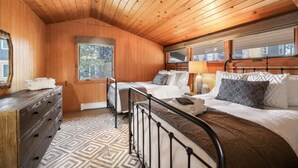 A cozy bedroom with wooden walls and ceiling, a large window, a bed with multiple pillows, a dresser, and a patterned rug.