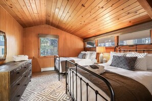 A cozy bedroom with wooden walls and ceiling, a large window, a bed with multiple pillows, a dresser, and a patterned rug.