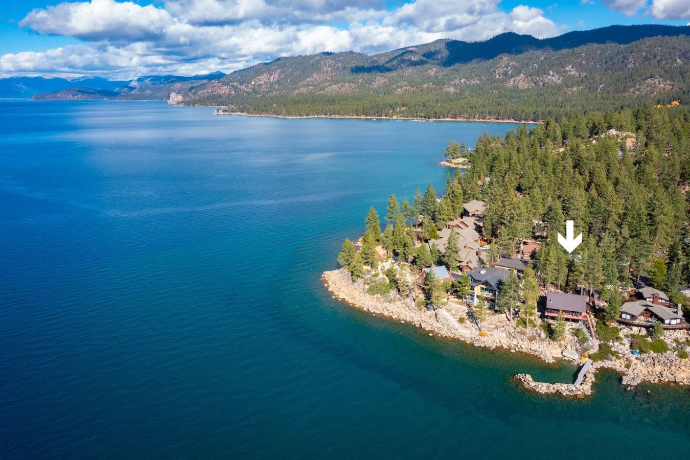 Vue aérienne d’une propriété au bord d’un lac, comprenant plusieurs bâtiments, entourée d’une forêt dense et avec des montagnes en arrière-plan.