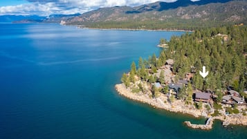 Vue aérienne d’une propriété au bord d’un lac, comprenant plusieurs bâtiments, entourée d’une forêt dense et avec des montagnes en arrière-plan.