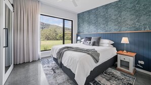Ein modernes Schlafzimmer mit einem großen Bett, einem Nachttisch, einer Lampe und Blick auf eine grüne Landschaft durch ein großes Fenster.