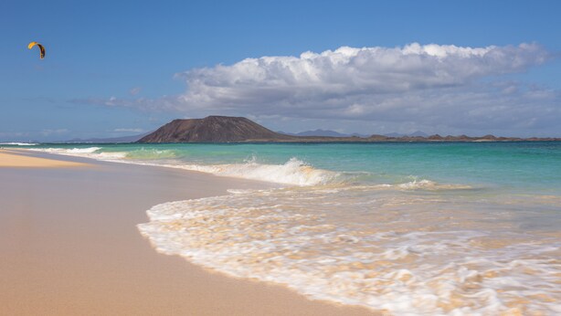 Een strand met helder turquoise water, een zandstrand en een berg op de achtergrond.