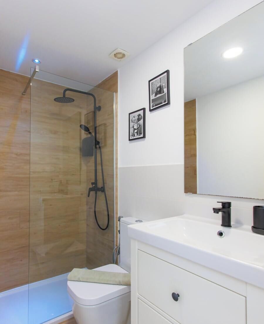 Un baño moderno con ducha envuelta en vidrio, un lavamanos blanco con grifo negro y dos cuadros enmarcados en la pared.