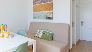 A living room with a beige sofa, a white table with breakfast items, and three wall art pieces depicting clouds, sand dunes, and a landscape.