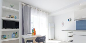 En moderne stue med et fladskærms-tv, et spisebord med stole, et køkkenområde med et køleskab og et vindue med gardiner.