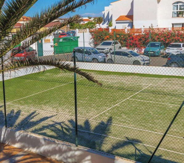 Área de estacionamento cercada, com vários carros, uma palmeira e um prédio com fachada branca ao fundo.