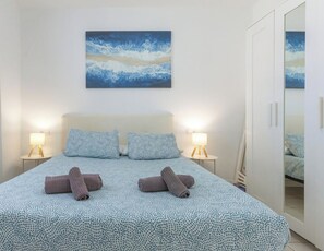 Un dormitorio con una cama, dos almohadas, una lámpara de noche, una toalla enrollada y un cuadro en la pared.