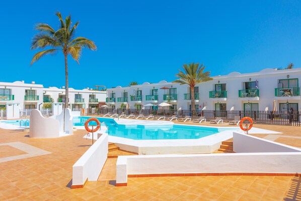 A pool area with a clear blue sky, palm trees, and a row of white buildings with balconies.