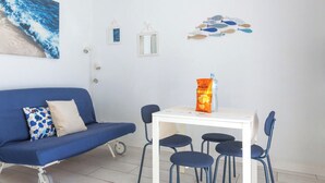 Une pièce avec un canapé bleu, une table blanche et quatre chaises, un tableau représentant l’océan et un élément décoratif mural représentant des poissons.