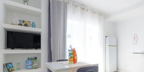 A hotel room with a flat-screen TV, a table with a bottle of water and a bag of chips, a window with curtains, and a refrigerator.