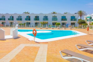 Área de piscina com espreguiçadeiras, um flutuador salva-vidas e um prédio com várias varandas.