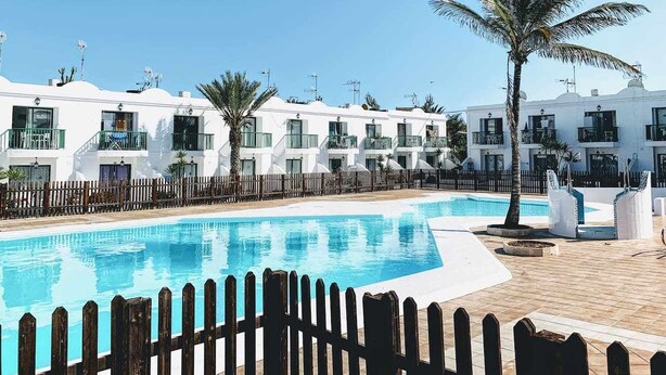 A pool area with a wooden fence, white buildings with balconies, and palm trees. - Desert Pool View & WiFi by iRent Fuerteventura (Corralejo Playa)
