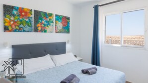 Une chambre à coucher avec un lit, une fenêtre donnant sur la plage et trois tableaux colorés accrochés au mur.