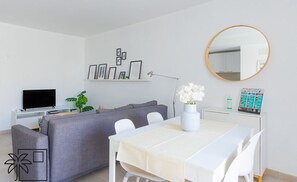 Ein modernes Wohnzimmer mit einem grauen Sofa, einem weißen Esstisch mit Stühlen, einem Flachbildfernseher, einer Pflanze, einem Spiegel und gerahmten Bildern an der Wand.