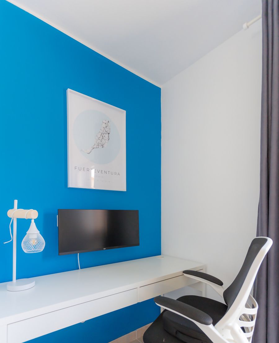 Une pièce avec un mur d’accent bleu, un téléviseur à écran plat fixé sur un bureau blanc, un fauteuil de bureau noir et une œuvre d’art encadrée au-dessus du bureau.