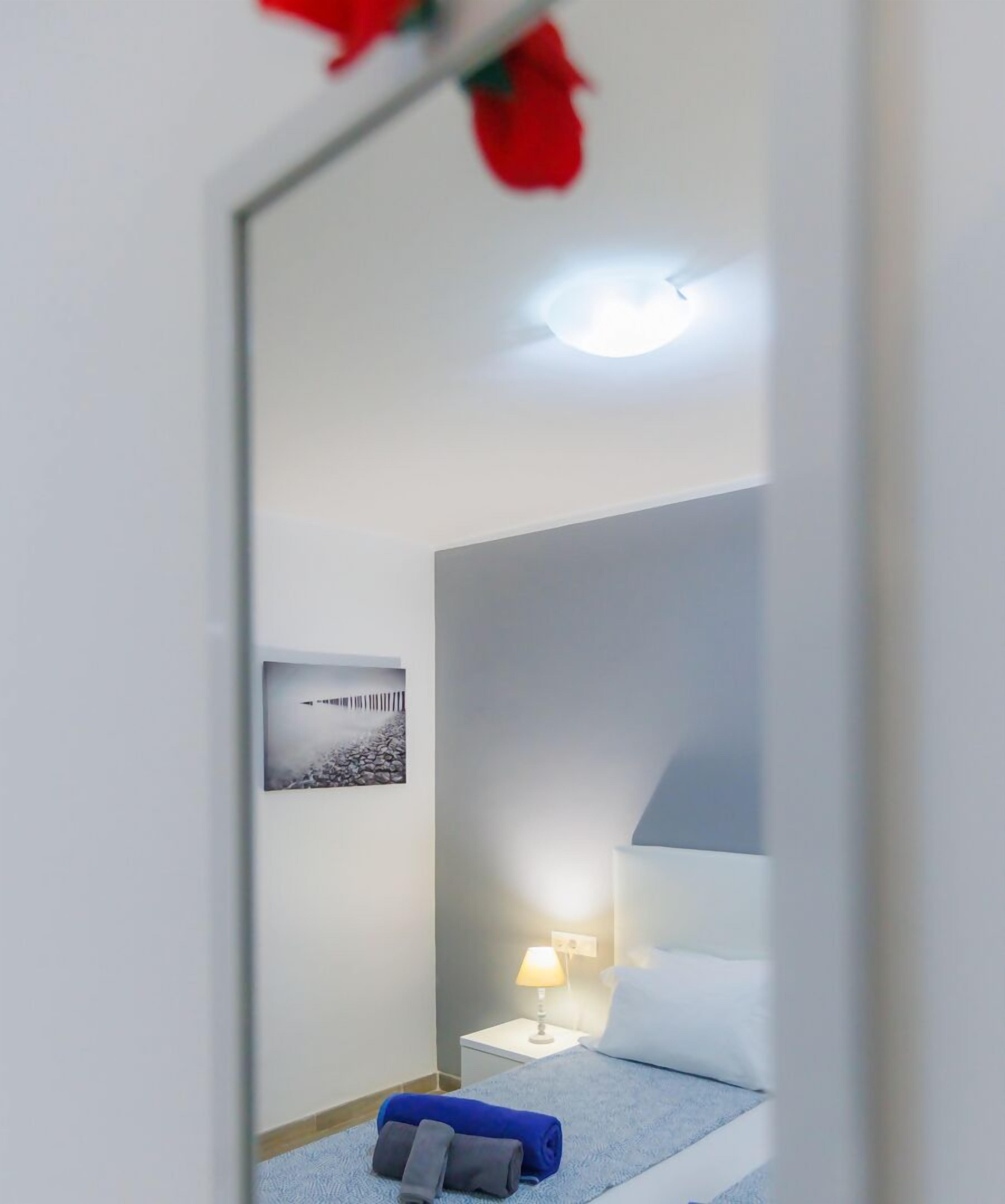 A bedroom with a bed, a nightstand, a lamp, a picture on the wall, and a mirror reflecting the room.