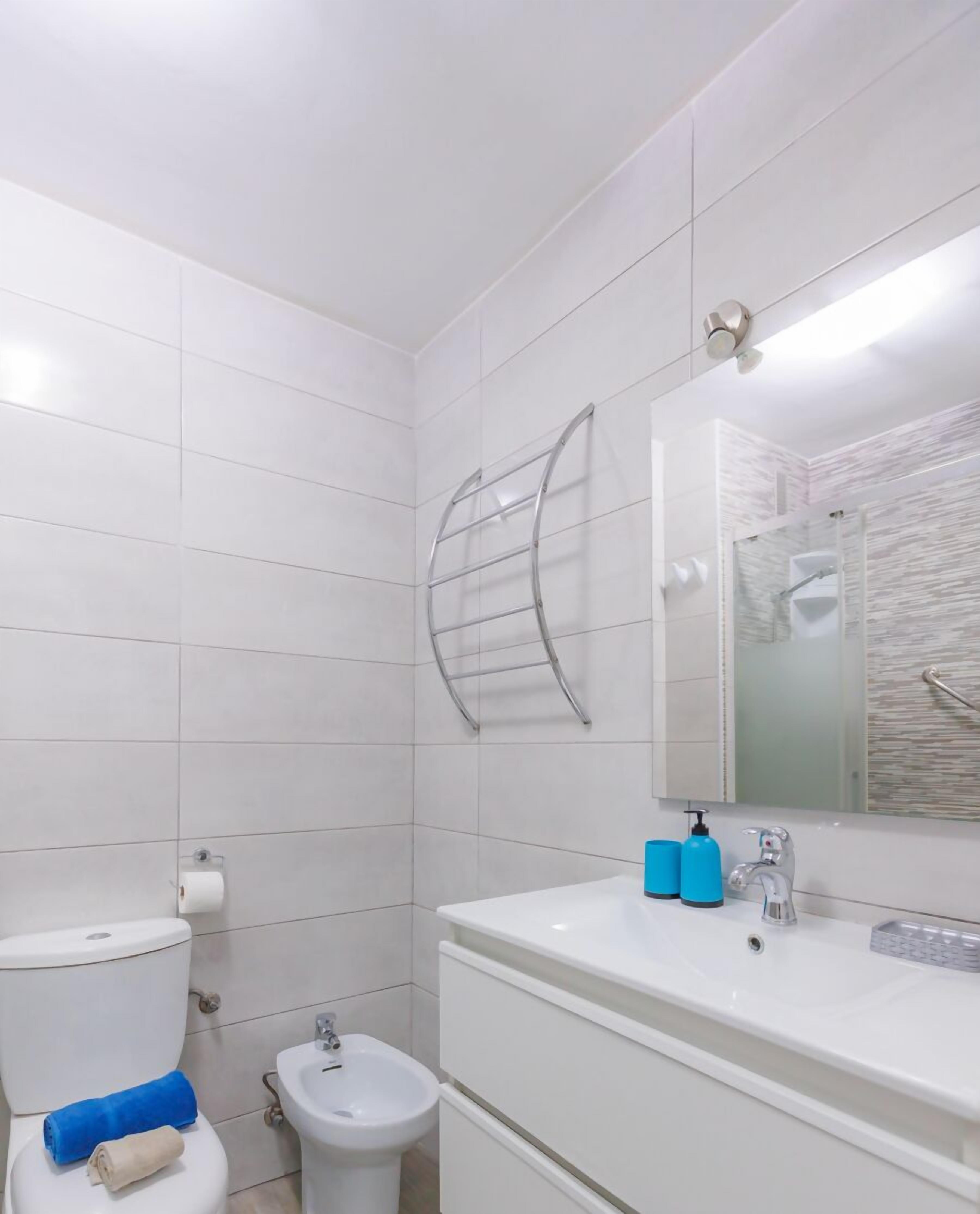 Ein modernes Badezimmer mit weißen Fliesen, Toilette, Bidet, Waschtisch mit Spiegel und Handtuchhalter.