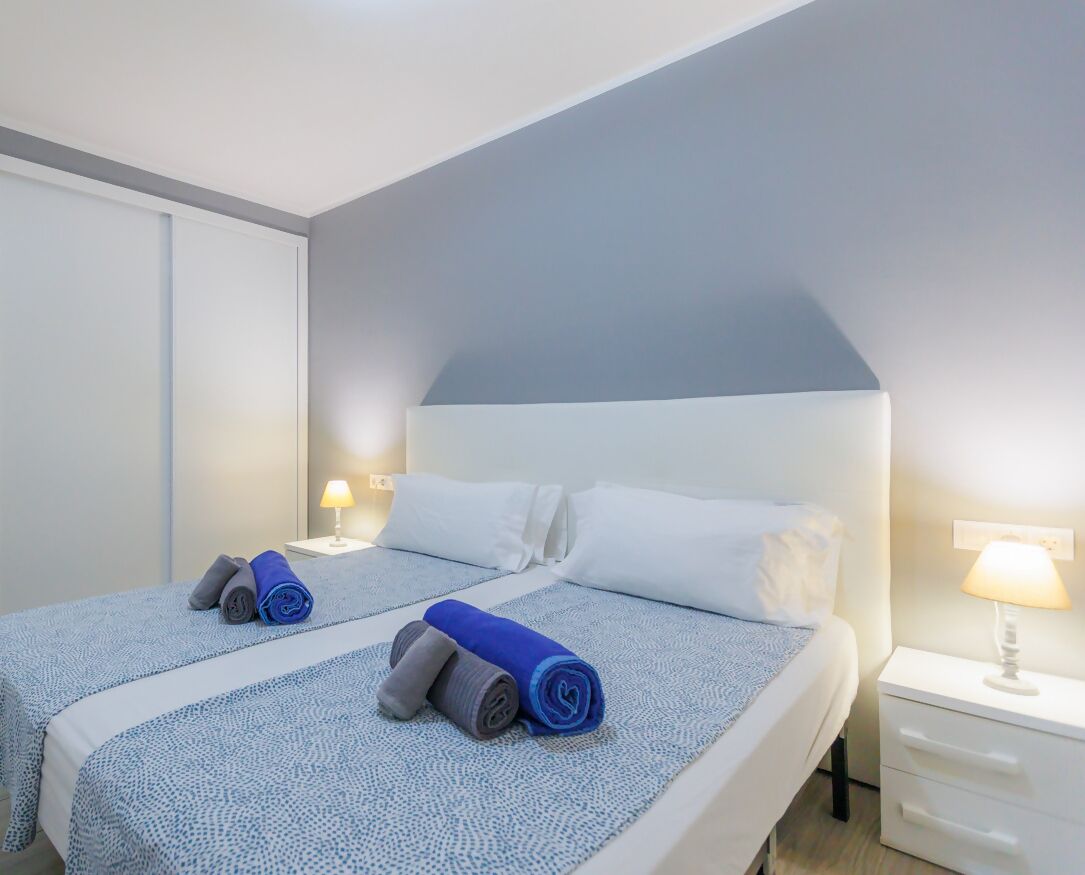 A hotel room with a double bed, two pillows, a nightstand with a lamp, and a white dresser.