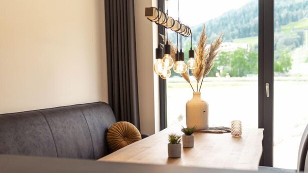 Un comedor moderno con una mesa de madera, un sofá gris oscuro y vistas a zonas verdes a través de amplios ventanales.