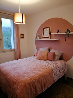 A bedroom with a bed, a hanging light fixture, a window with curtains, and a wall-mounted shelf with decorative items.