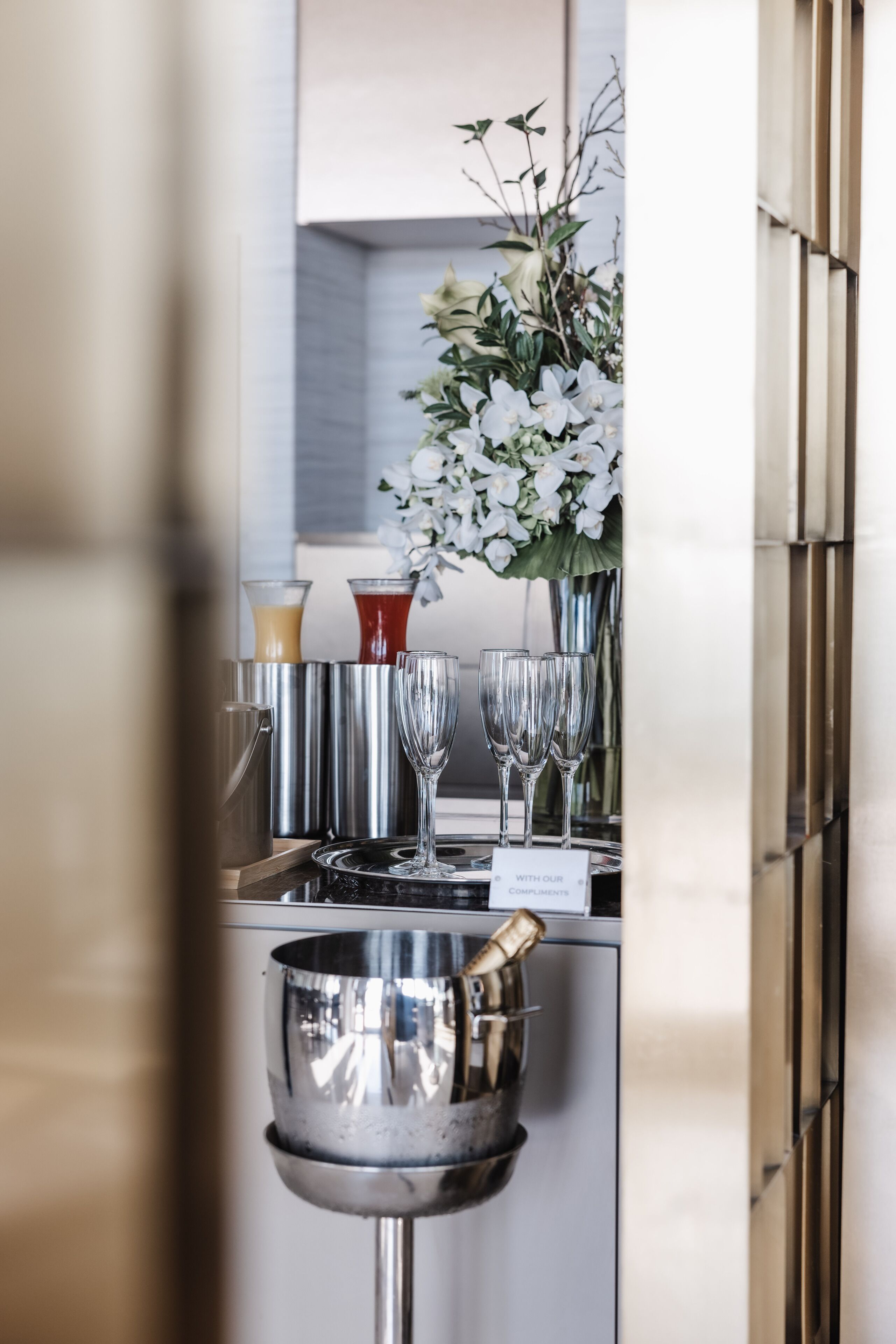 Un espace bar doté d’un comptoir en acier inoxydable, comprenant un shaker à cocktails, des verres et un arrangement floral.