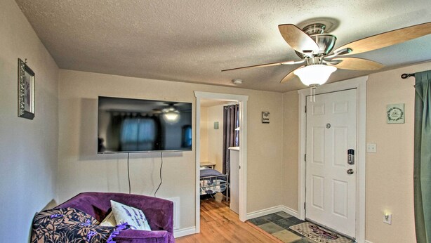 A living room with a purple armchair, a ceiling fan, a flat-screen TV, and a hallway leading to a bedroom with a bed and a door. - Lovely Tacoma Cottage w/ Fire Pit, Near Dtwn! (Tacoma)
