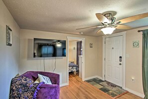 A living room with a purple armchair, a ceiling fan, a flat-screen TV, and a hallway leading to a bedroom with a bed and a door. - Lovely Tacoma Cottage w/ Fire Pit, Near Dtwn! (Tacoma)